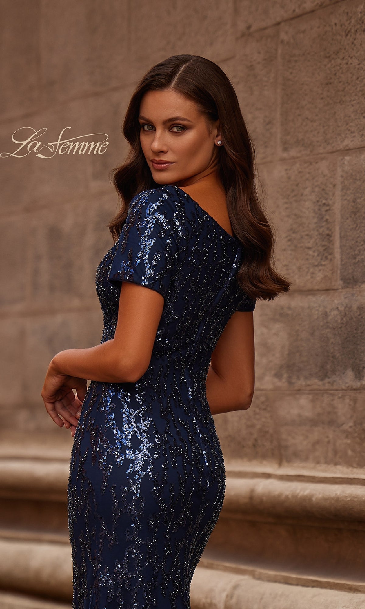 A woman with long brown hair models the La Femme 31787 Long Navy Sequin Formal Dress, standing against a stone wall and glancing over her shoulder.